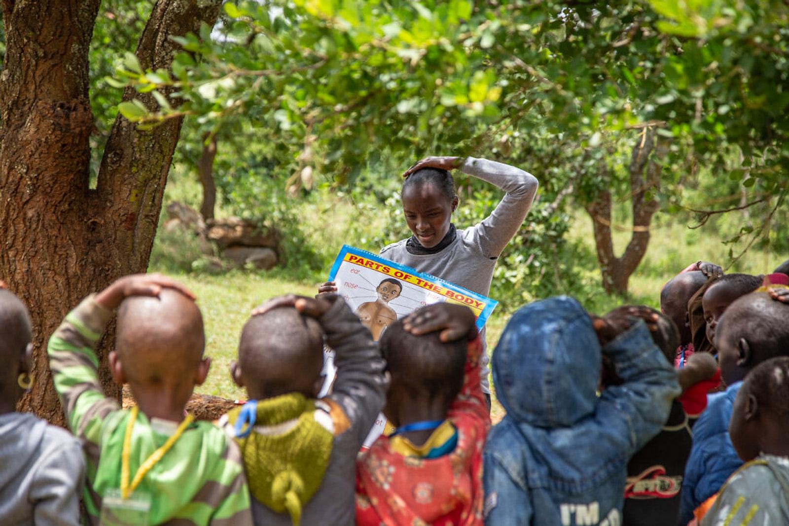 A young facilitator teaching children under a tree, holding up a learning chart